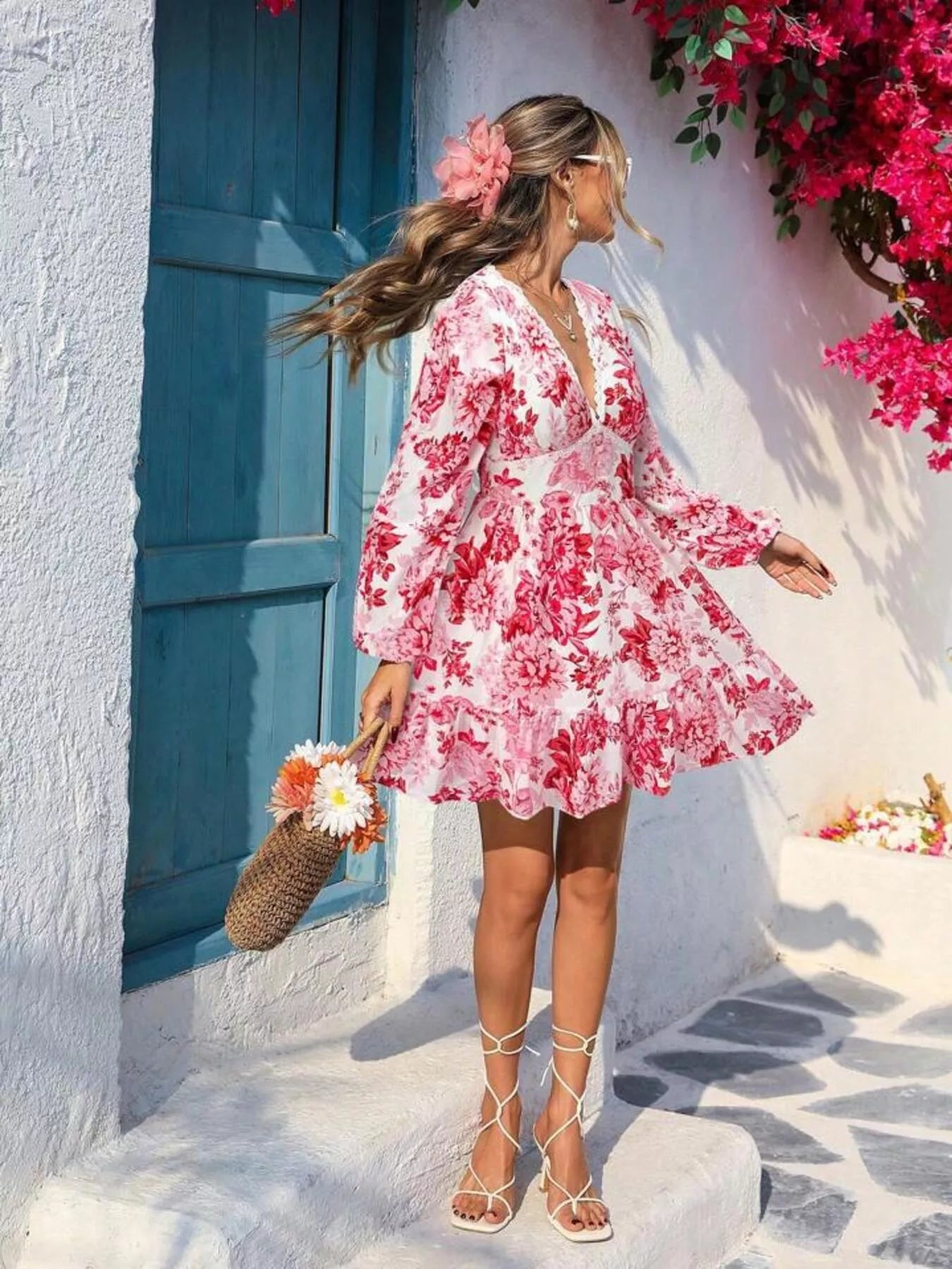Woman in a pink floral dress standing in front of a white wall with a blue door and pink flowers.