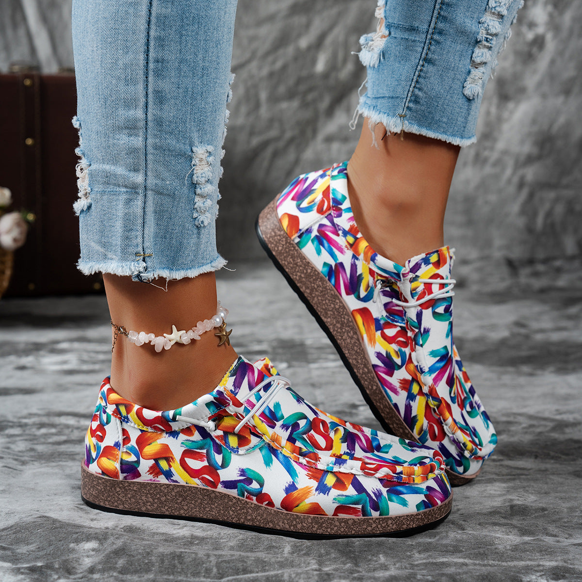 Colorful slip-on shoes with text pattern worn with ripped jeans on a gray surface.