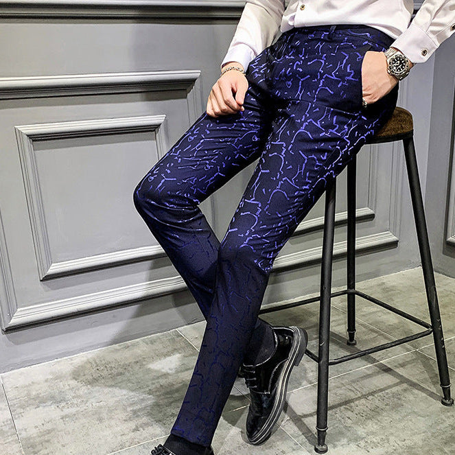 Person wearing blue patterned pants and black shoes sitting on a stool against a gray wall.