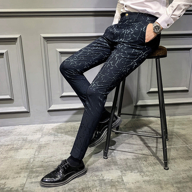 Person wearing dark patterned pants and black shoes sitting on a stool against a gray wall.