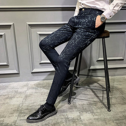Person wearing dark patterned pants and black shoes sitting on a stool against a gray wall.