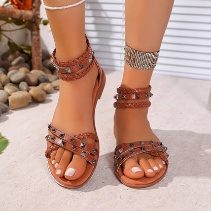 Brown strappy sandals with studs worn by a person on a sandy surface.