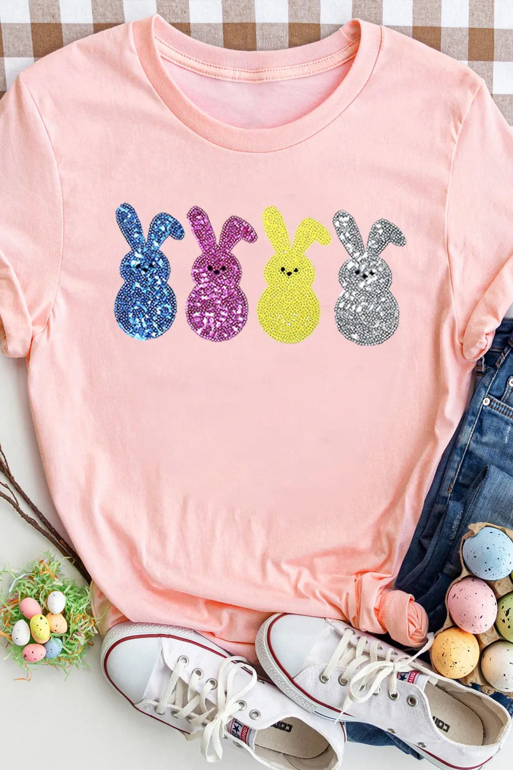 Pink t-shirt with colorful sequin bunnies, white sneakers, and Easter eggs on a checkered tablecloth.