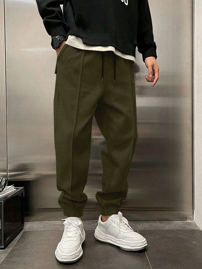 Person wearing a black sweatshirt, green pants, and white sneakers in front of a metallic wall.