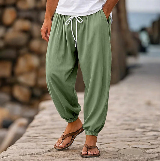 Person wearing green baggy pants and brown sandals standing on a stone path with a wooden log pile in the background.