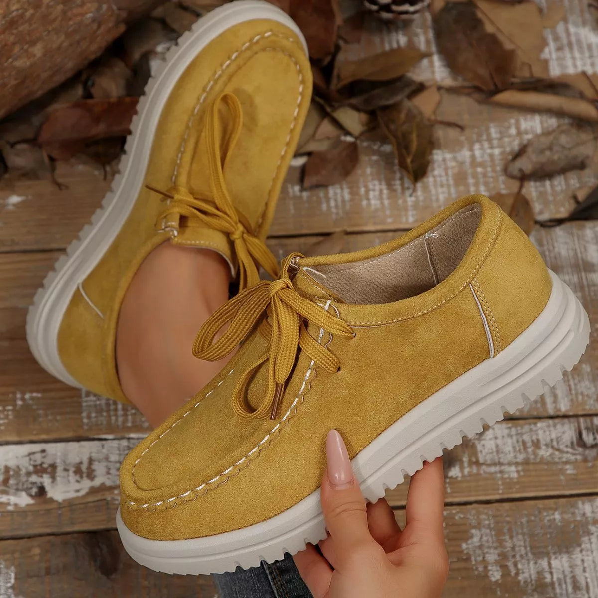Yellow suede shoes with white soles held by a hand on a wooden floor with leaves.