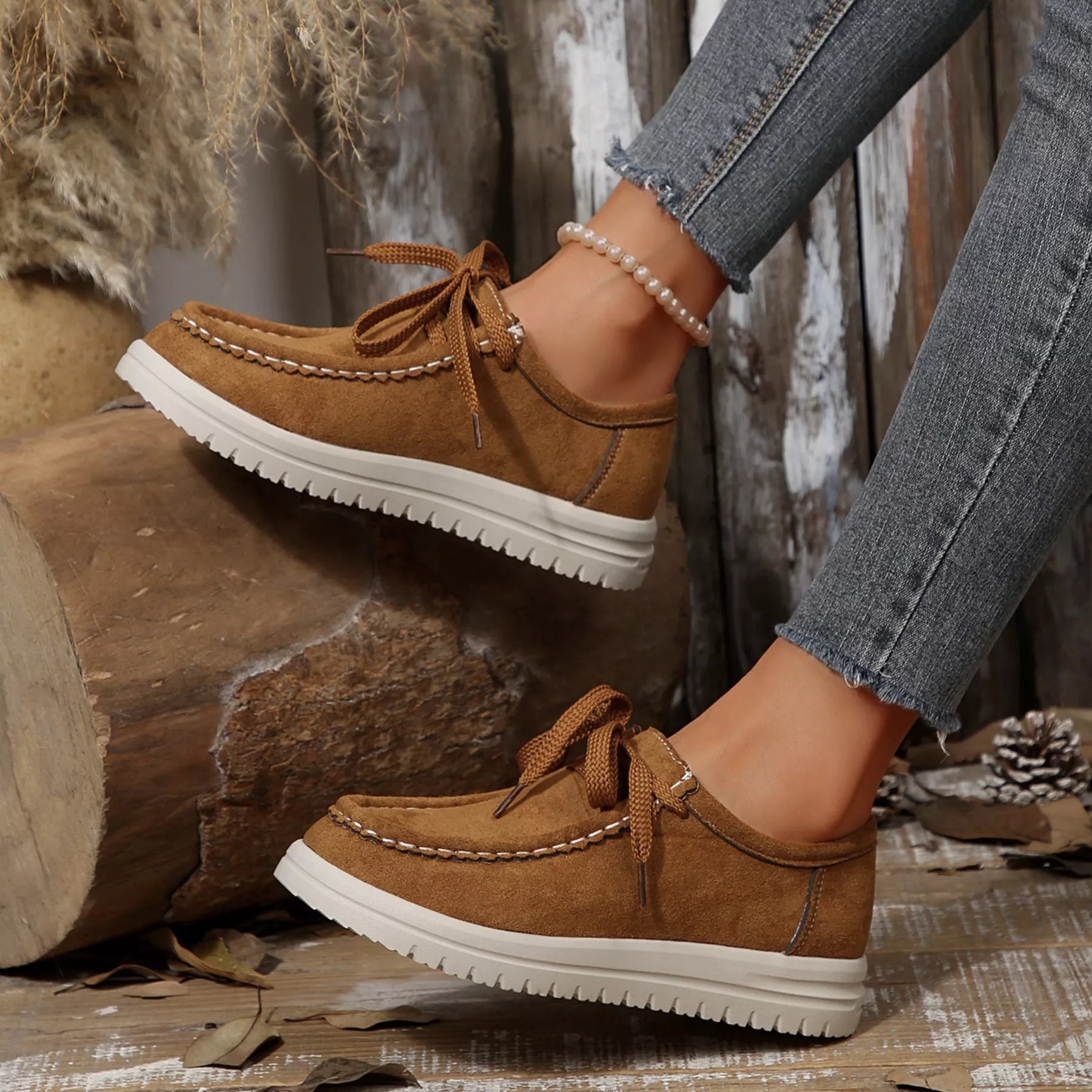 Brown suede shoes with white soles worn by a person sitting against a rustic wooden background.