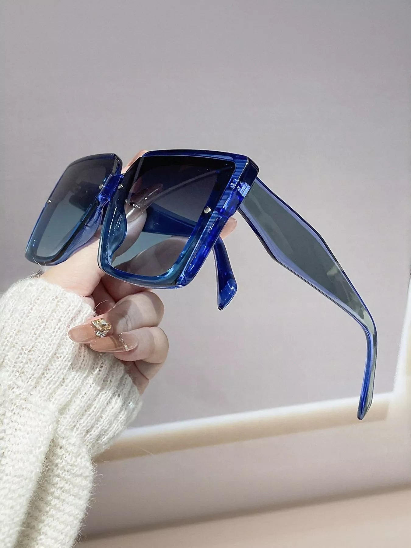 Blue sunglasses held by a hand wearing a white sweater against a neutral background