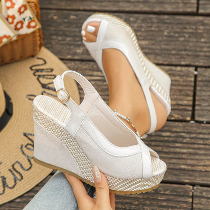 White wedge sandals with woven sole held by a hand on a wooden floor.