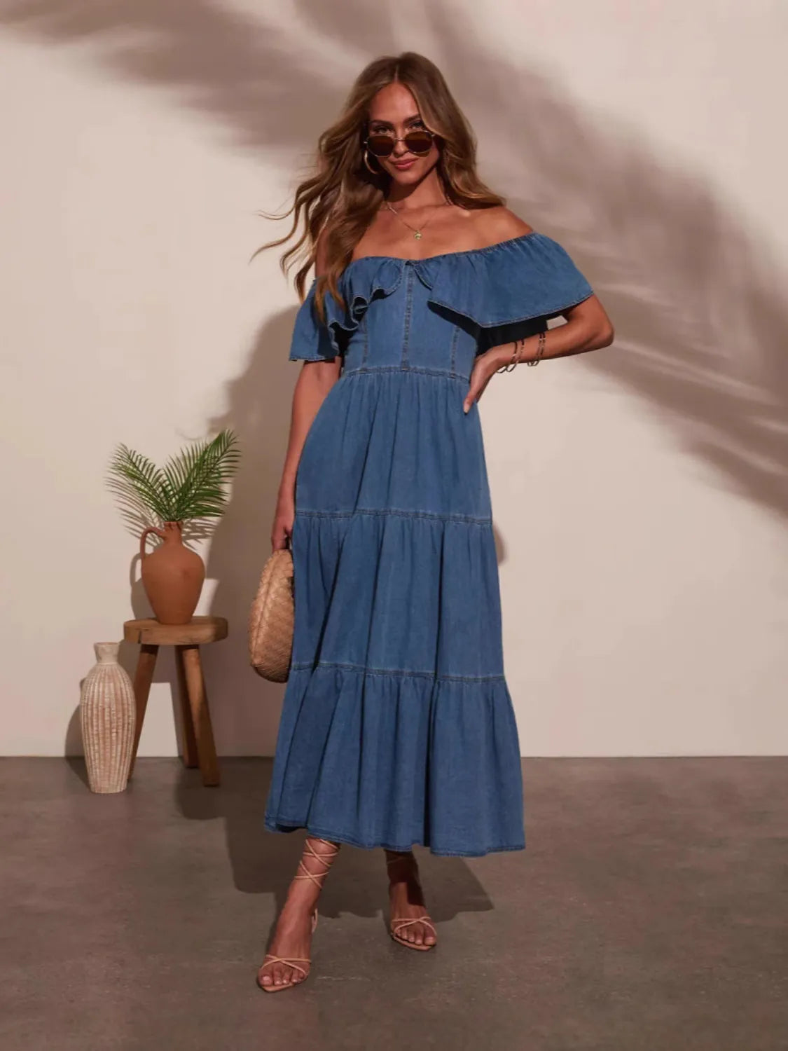 Woman wearing a blue off-shoulder dress standing against a plain wall with a plant and small table in the background.