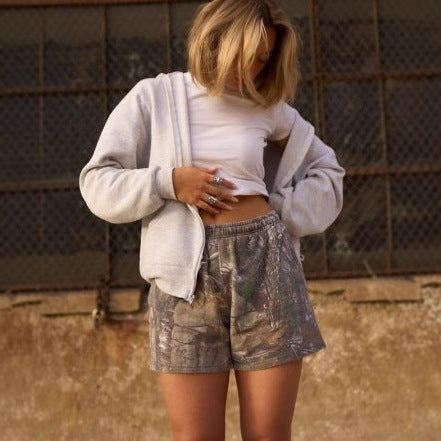 Person wearing a white crop top, gray sweatshirt, and camouflage shorts in an outdoor setting.