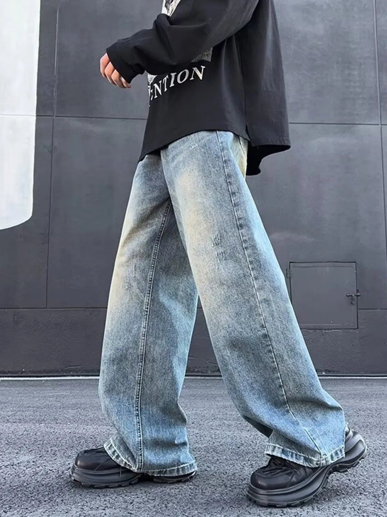 Person wearing oversized light blue jeans and black shoes against a gray wall.