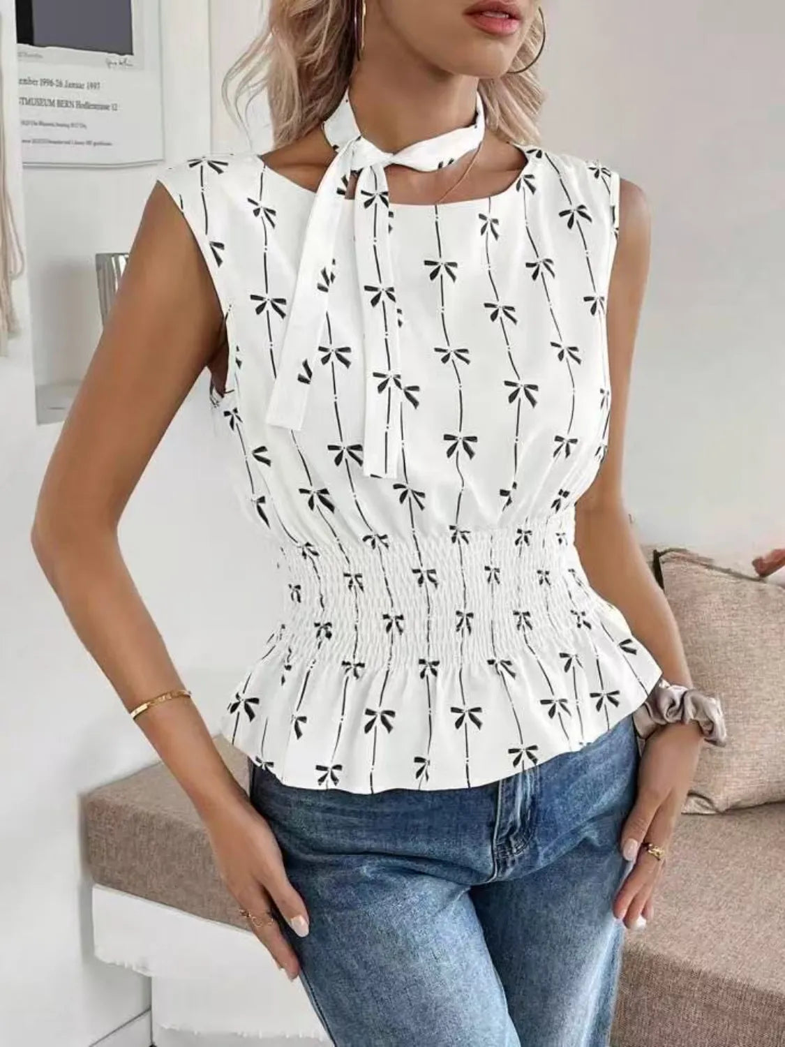 Woman wearing a white sleeveless top with black bow pattern and blue jeans indoors.
