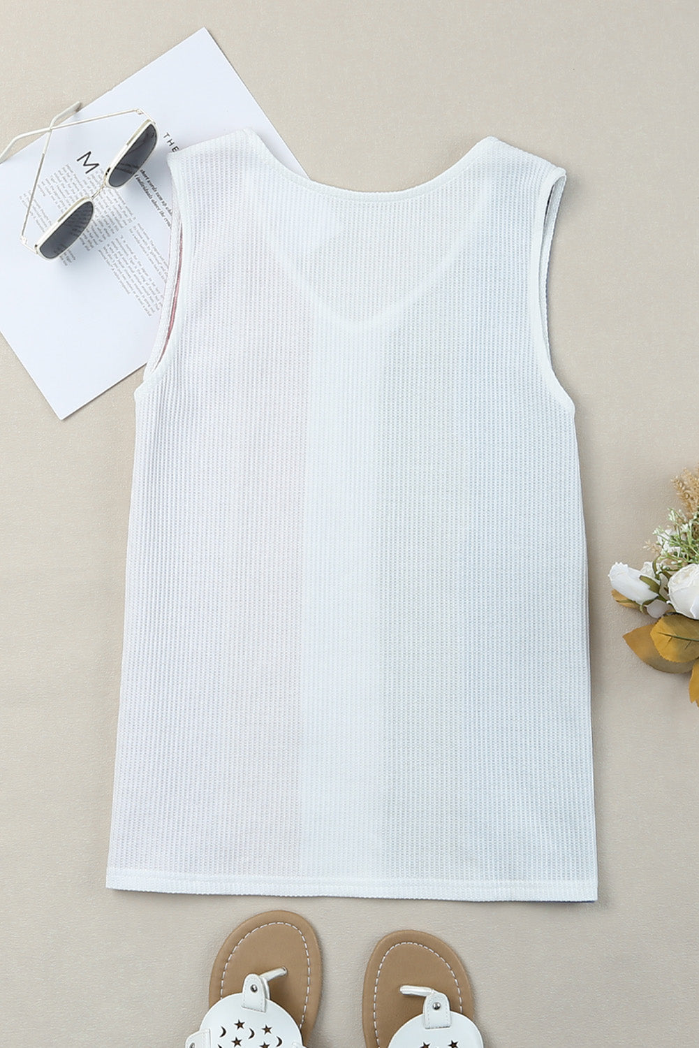 White ribbed tank top on a beige surface with sunglasses and sandals.