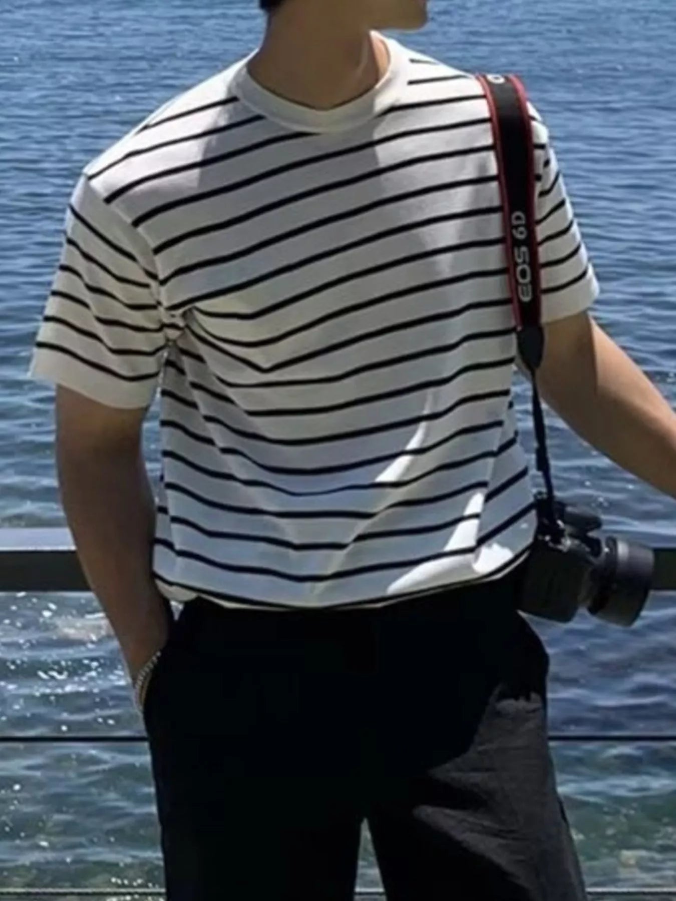 Person wearing a striped shirt with a camera around their neck, standing by water.