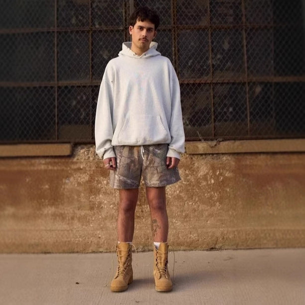 Person wearing a light gray hoodie, patterned shorts, and tan boots standing against a textured wall.