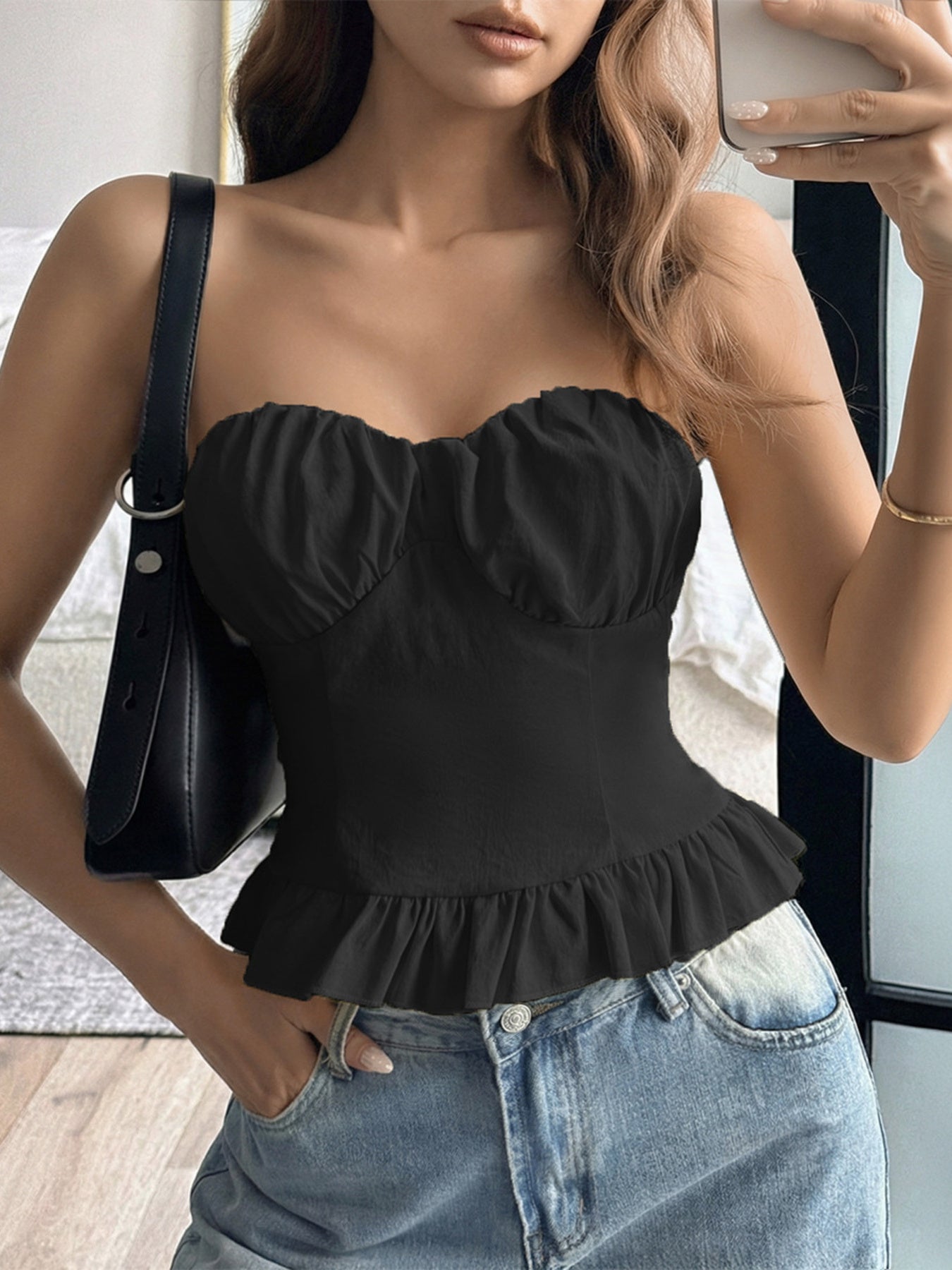 Woman wearing a black strapless top with ruffled details and blue jeans, holding a phone.