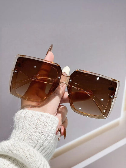 Hand holding a pair of sunglasses with brown lenses and gold frames against a neutral background