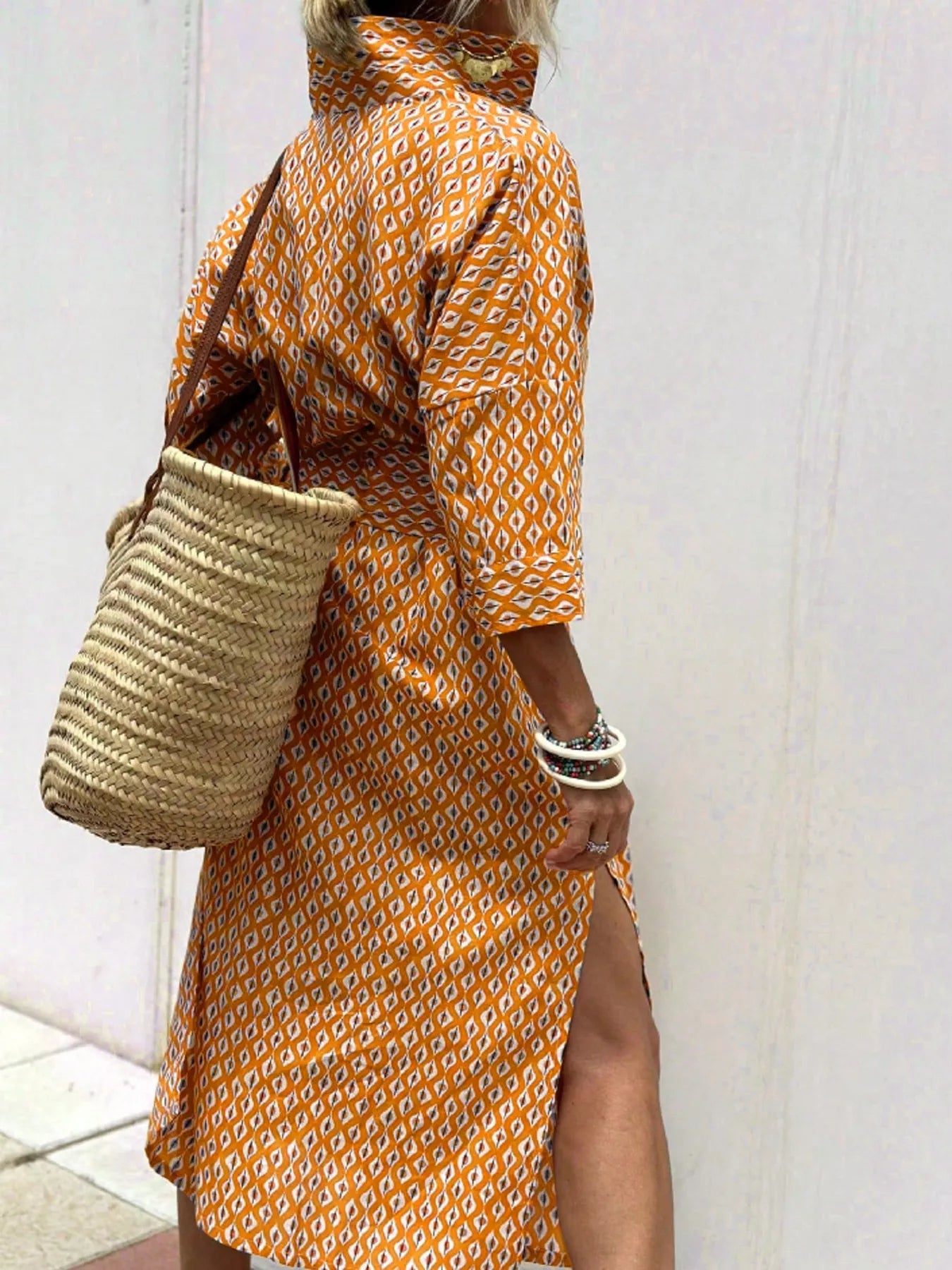 Person wearing an orange patterned dress holding a straw bag against a white wall.
