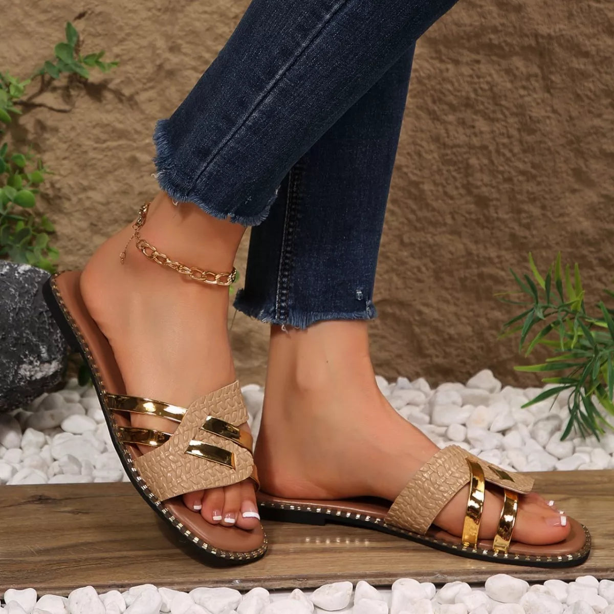 Beige sandals with gold straps worn by a person on a stone surface with plants in the background