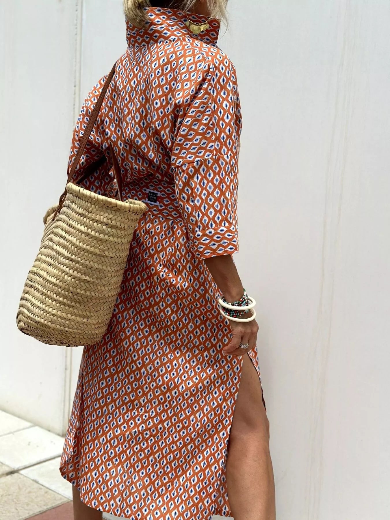 Person wearing a patterned dress holding a straw bag against a white wall.