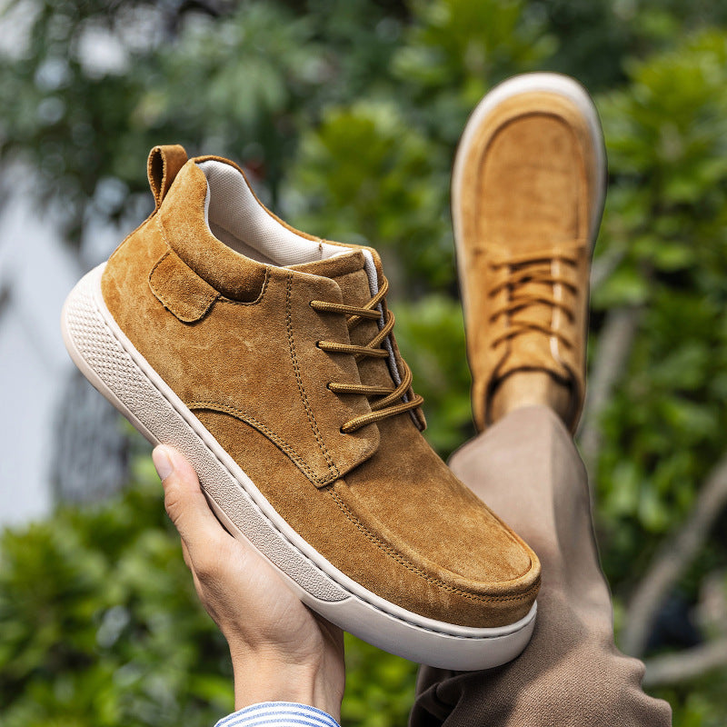 Brown suede shoes with white soles held by a hand against a blurred green outdoor background