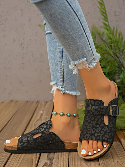 Person wearing black leopard print sandals with a wooden floor and plant in the background
