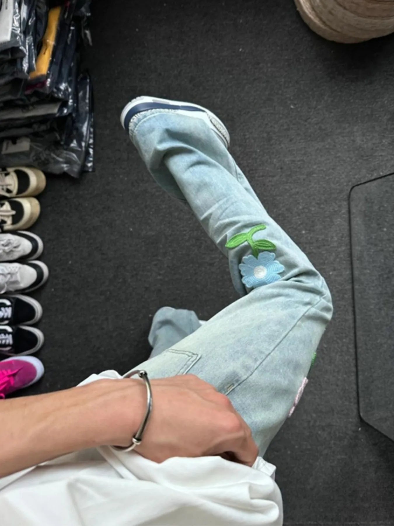 Person wearing light blue jeans with floral embroidery, sitting on a dark floor.