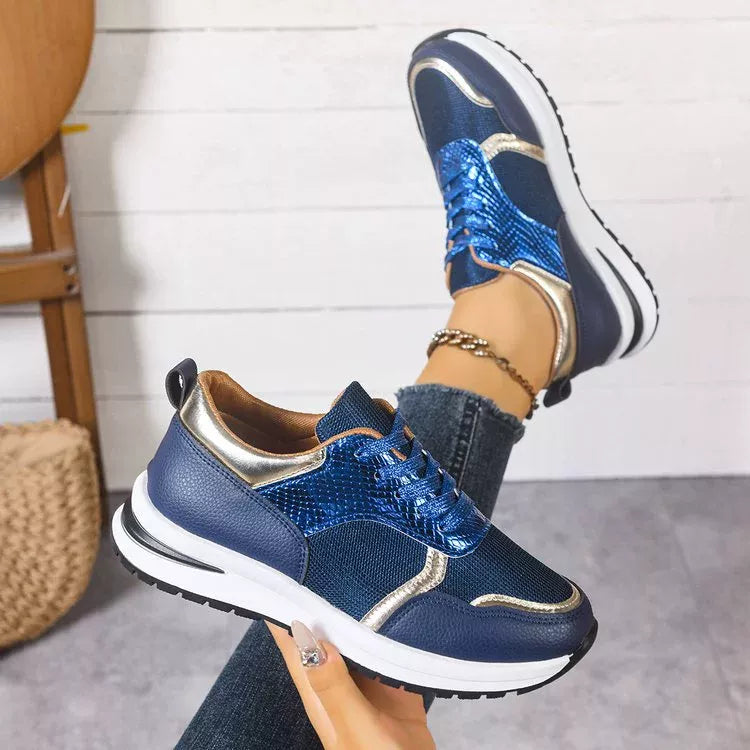 Blue sneakers with gold accents worn by a person against a white wall.