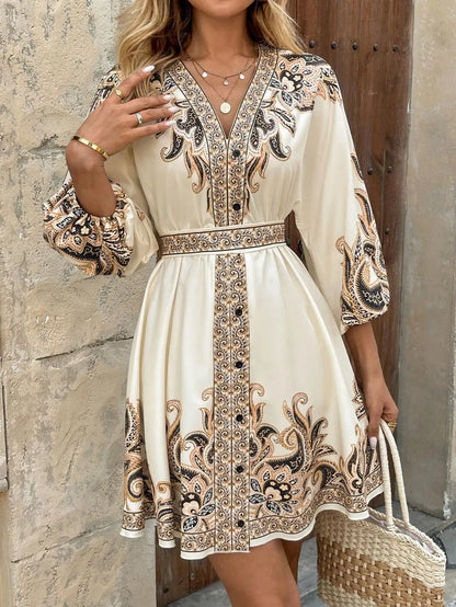 Woman wearing a cream-colored dress with intricate black patterns, standing against a stone wall.