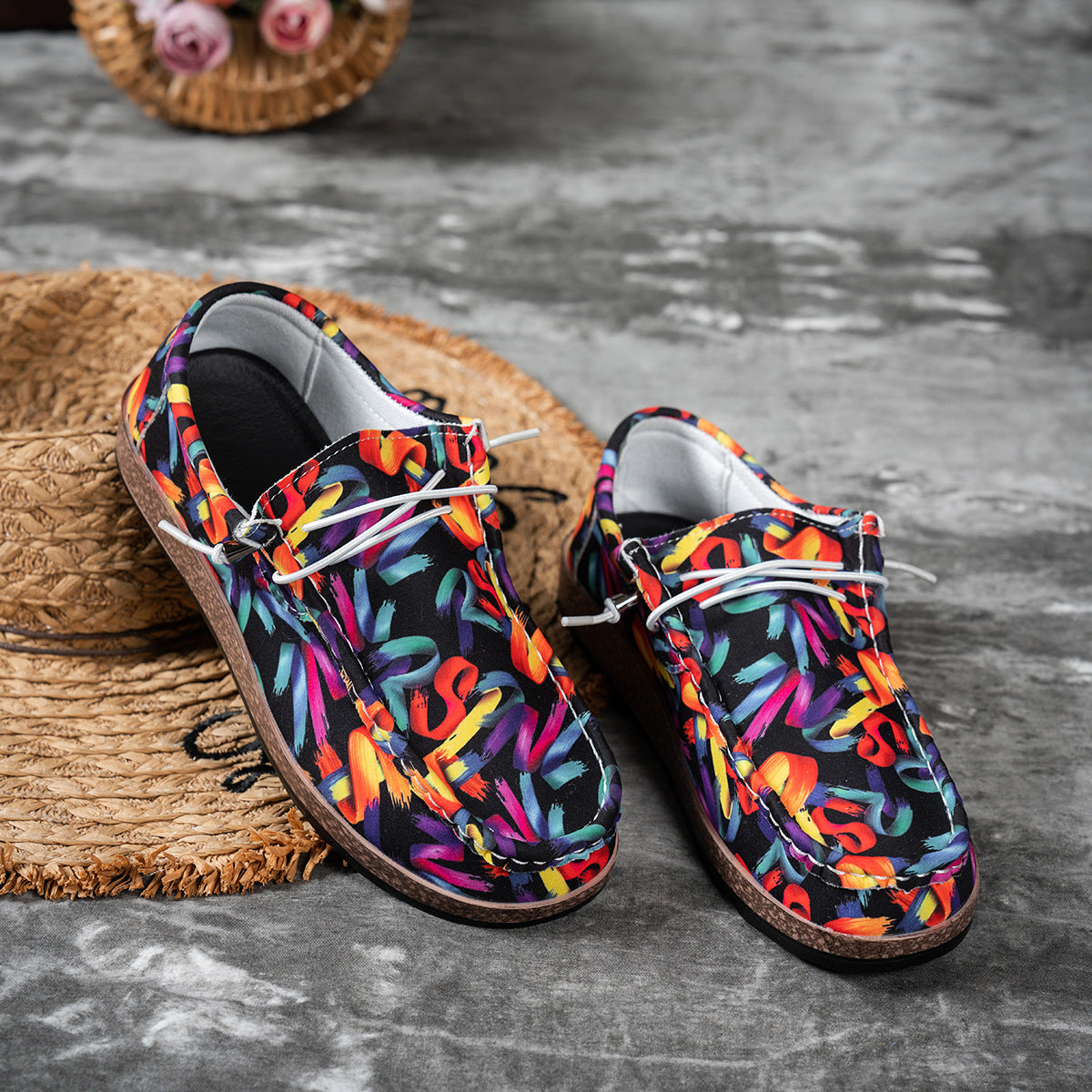 Colorful patterned shoes on a textured surface with a straw hat and flowers in the background.
