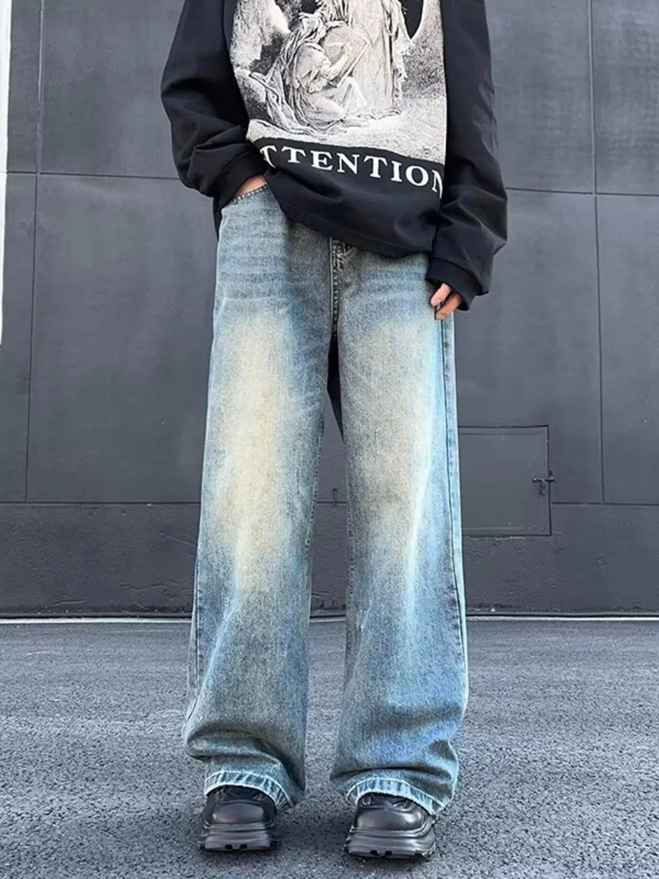 Person wearing a black sweatshirt with graphic design and light blue jeans against a gray wall.