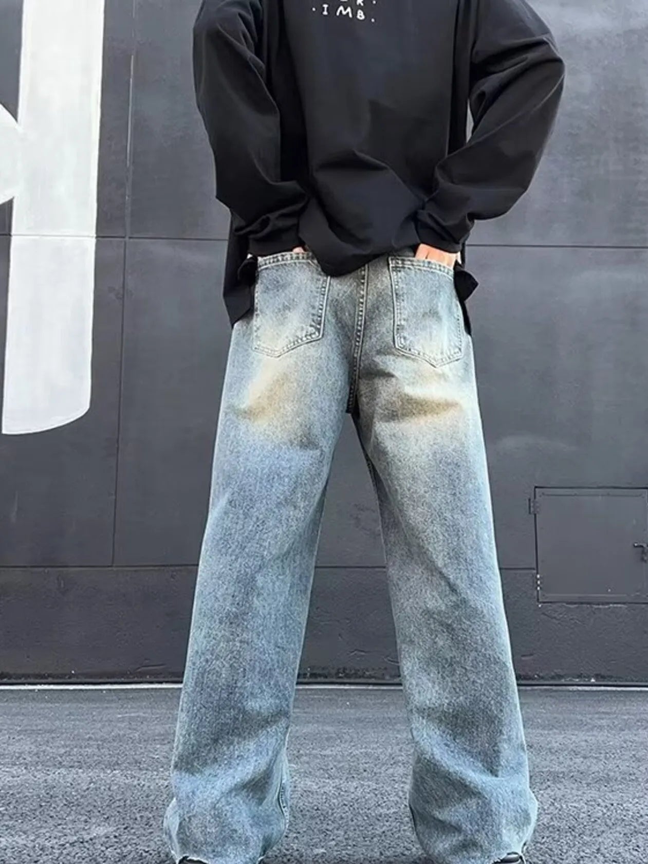 Person wearing a black sweatshirt and light blue jeans standing against a gray wall.