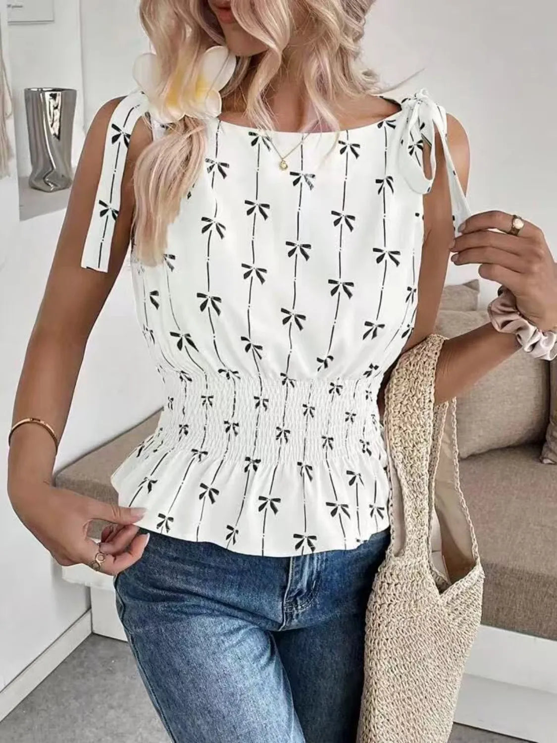 Woman wearing a white top with black patterns and blue jeans, holding a beige bag indoors.