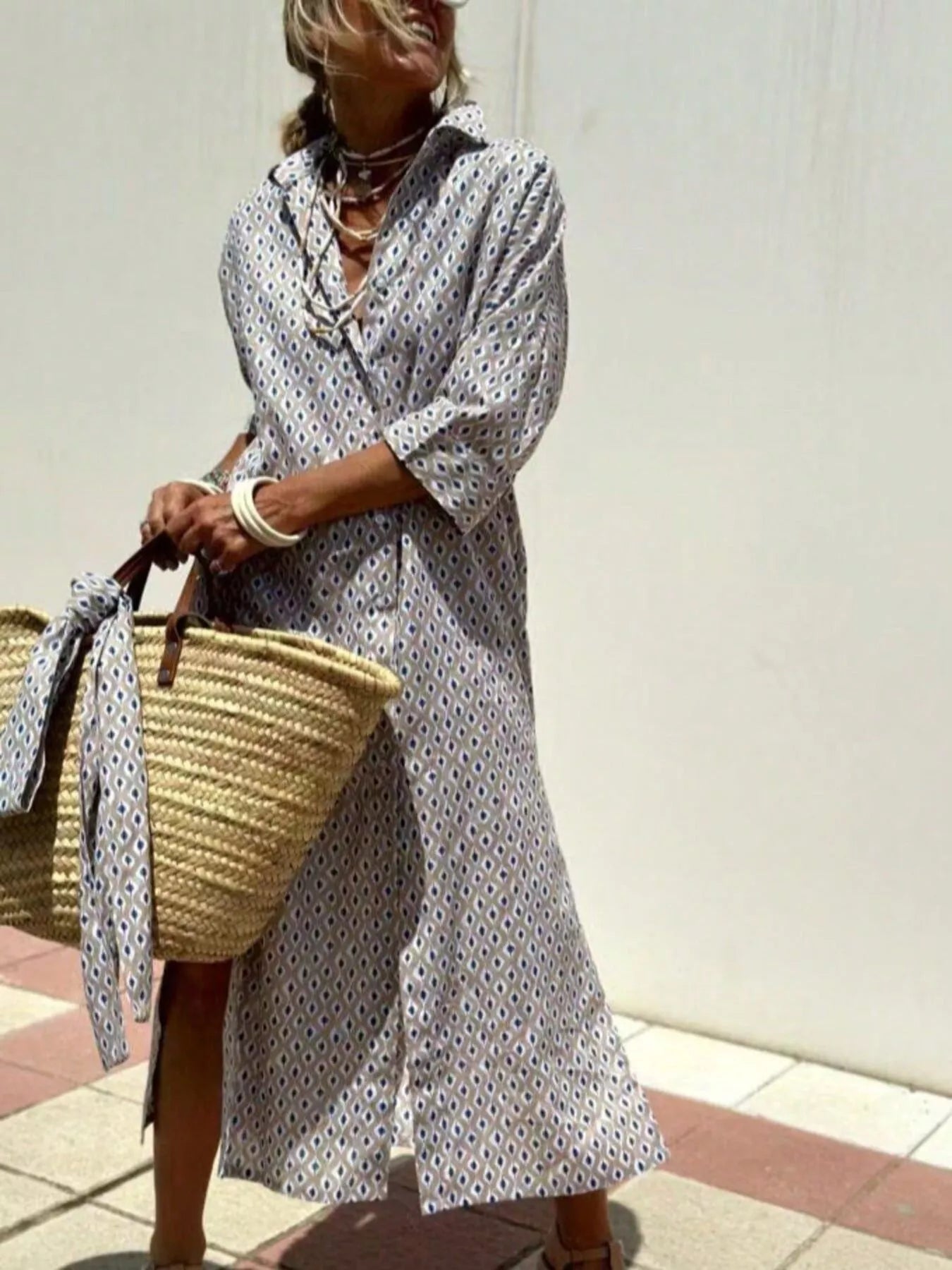 Woman in a white patterned dress holding a straw bag against a plain background
