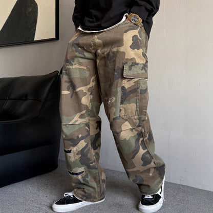 Person wearing camouflage pants and black sneakers in a room with a black bag and abstract art on the wall.