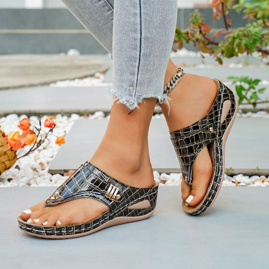 Black crocodile-patterned sandals worn with light blue jeans on a blurred outdoor background.