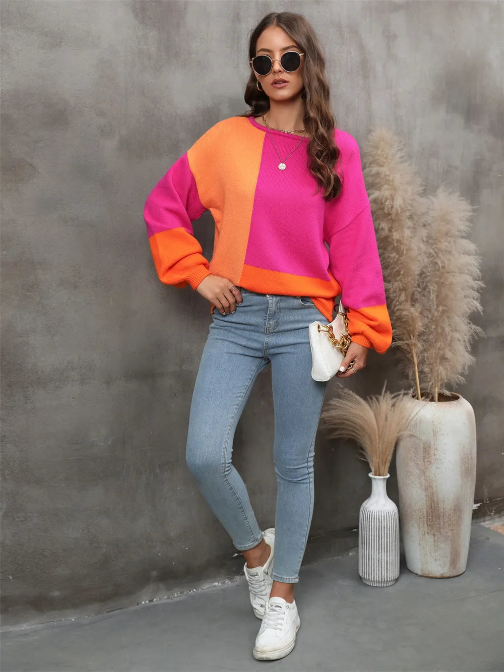 Woman wearing a colorful sweater and jeans standing against a gray wall with decorative plants.