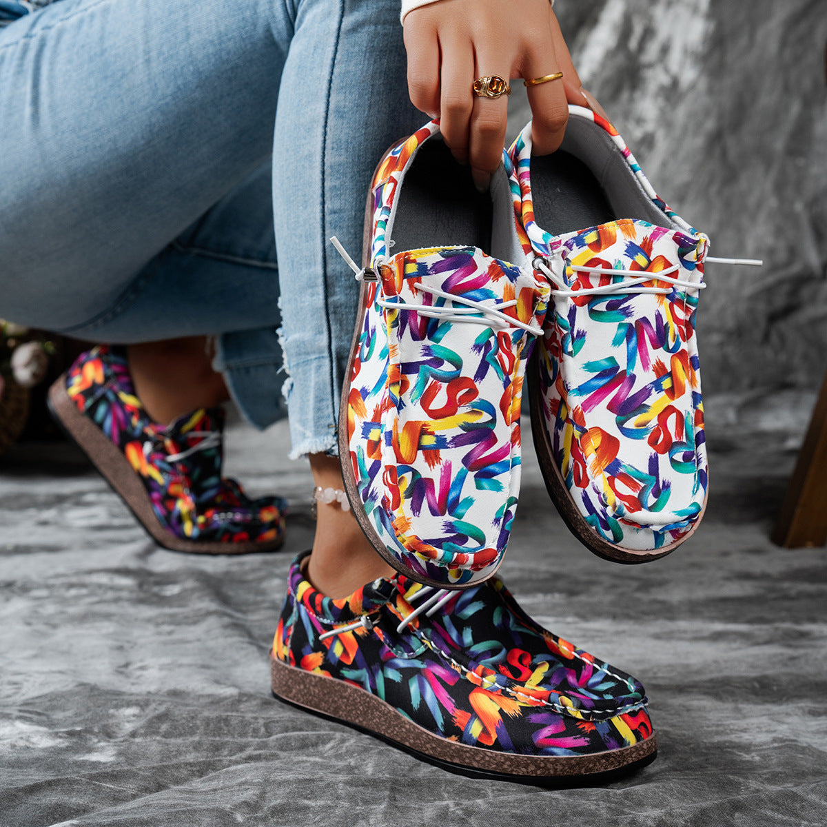 Colorful patterned shoes held by a person wearing jeans on a gray surface.