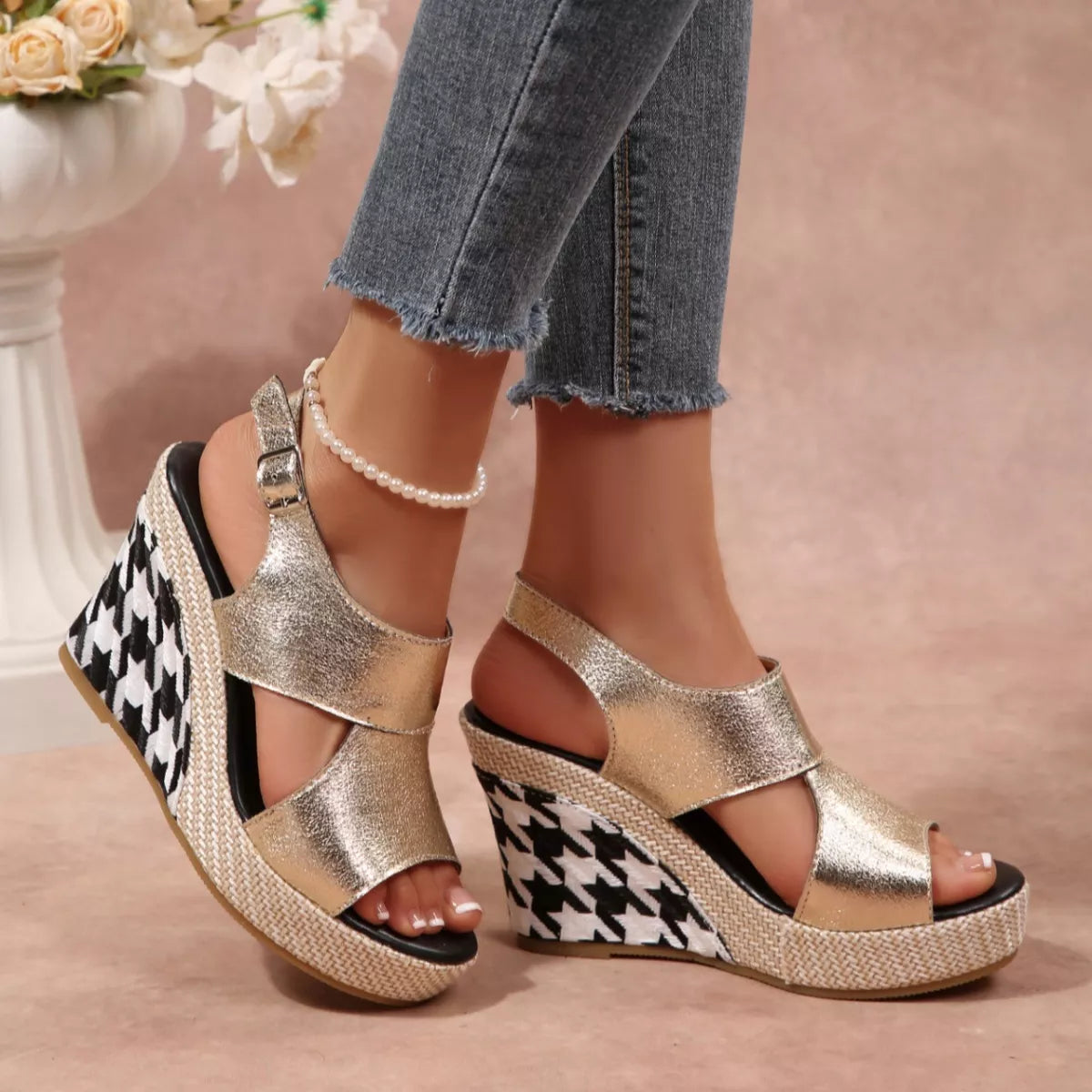 Gold wedge sandals with houndstooth pattern worn by a person, with a blurred floral background.